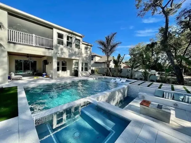 Luxury backyard pool with spa, patio loungers, and a two-story house under a clear blue sky