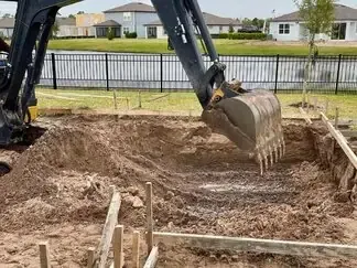 Excavator bucket digging a large dirt pit in a residential backyard with a fence and houses nearby