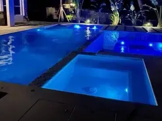 Blue-lit backyard swimming pool at night with an illuminated spa and dark patio.