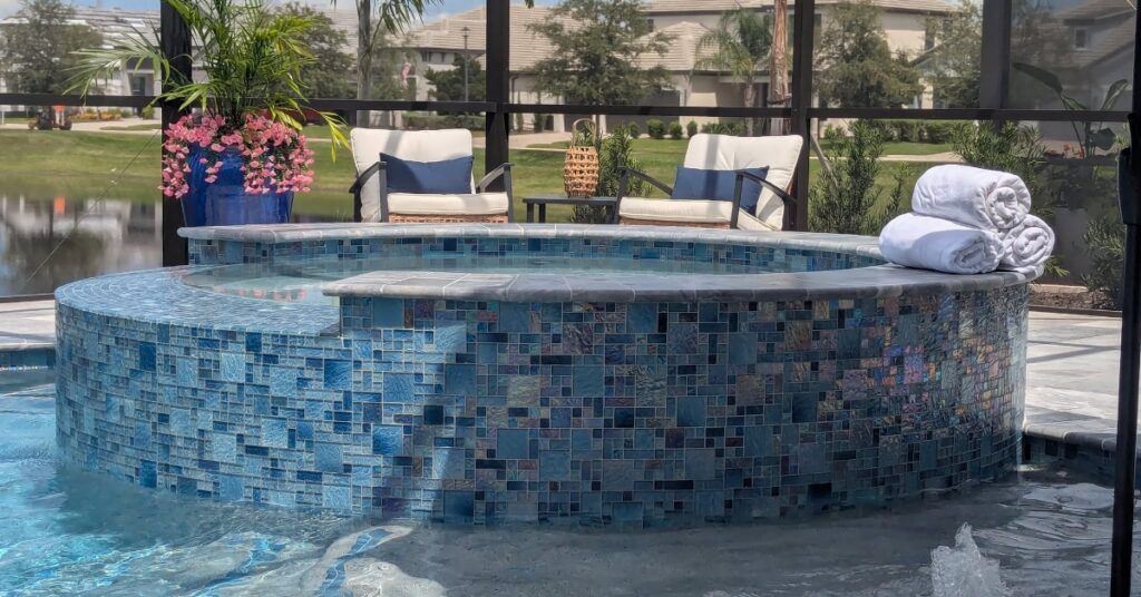 Blue mosaic poolside hot tub with white towels and lounge chairs in a fenced outdoor resort area