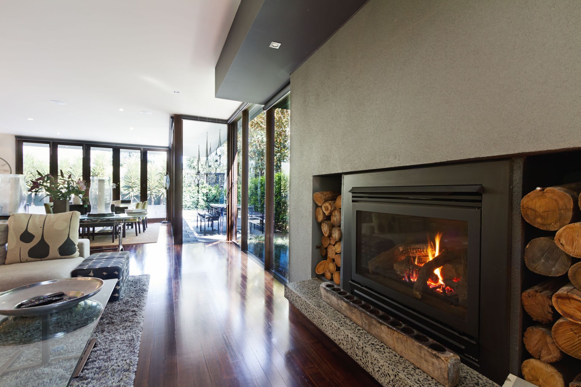 Living room with fireplace, firewood, and glass doors opening to patio. Wooden floors and neutral-toned furniture.