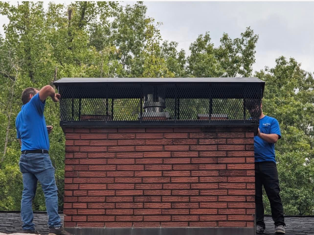 Two people installing a chimney cap on a brick chimney; green trees in the background.