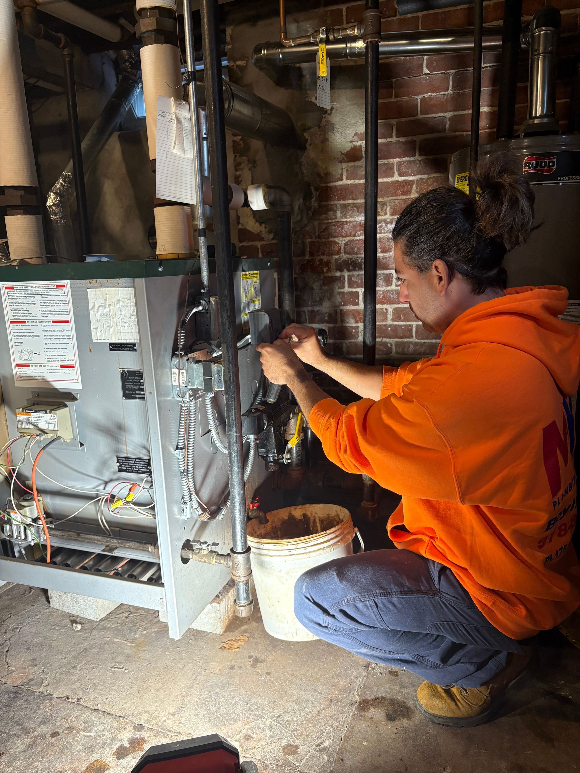 Man in orange hoodie working on a furnace in a basement, using tools.