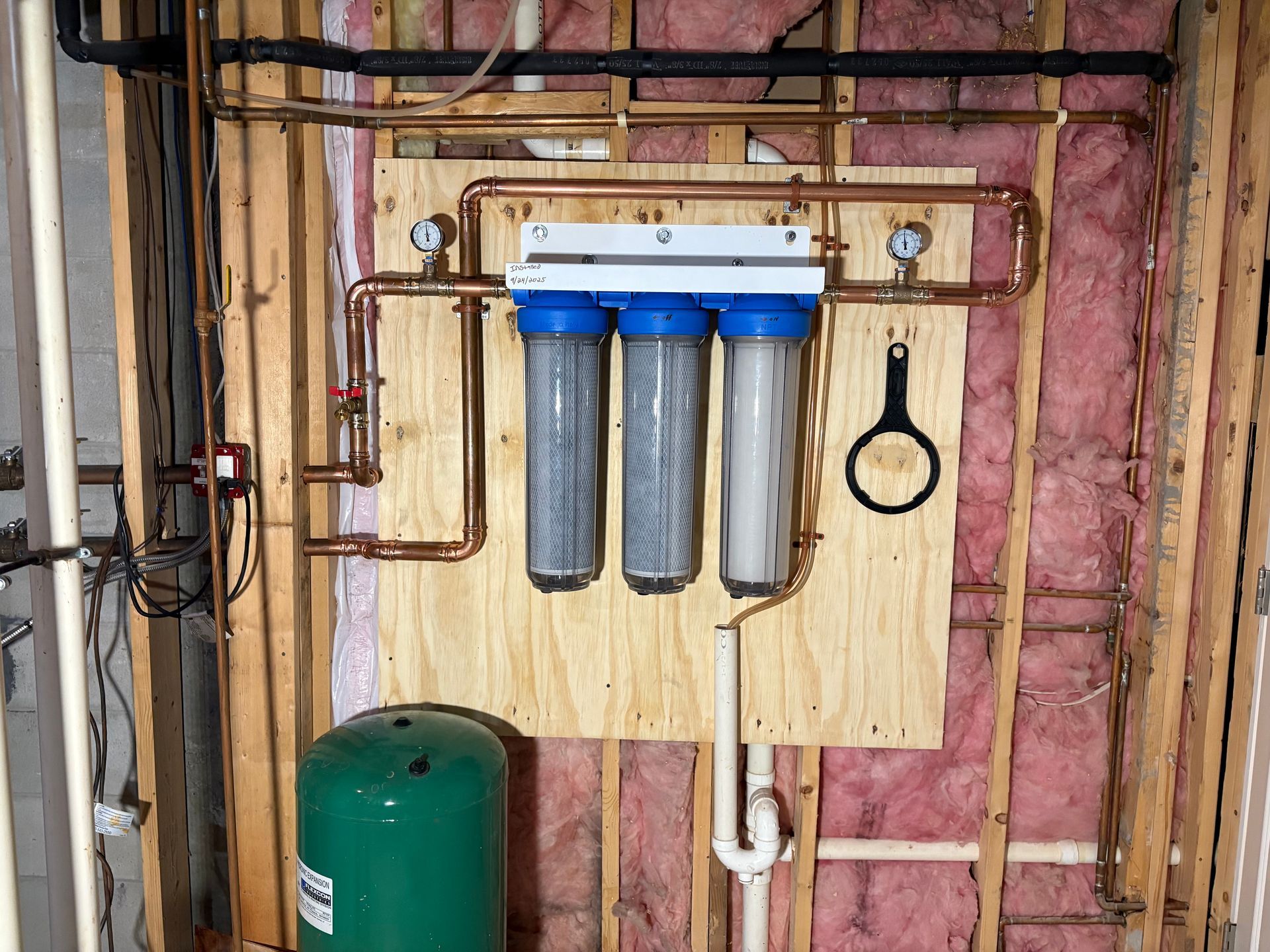 Water filtration system with three blue canisters mounted on plywood, copper piping, and pink insulation.
