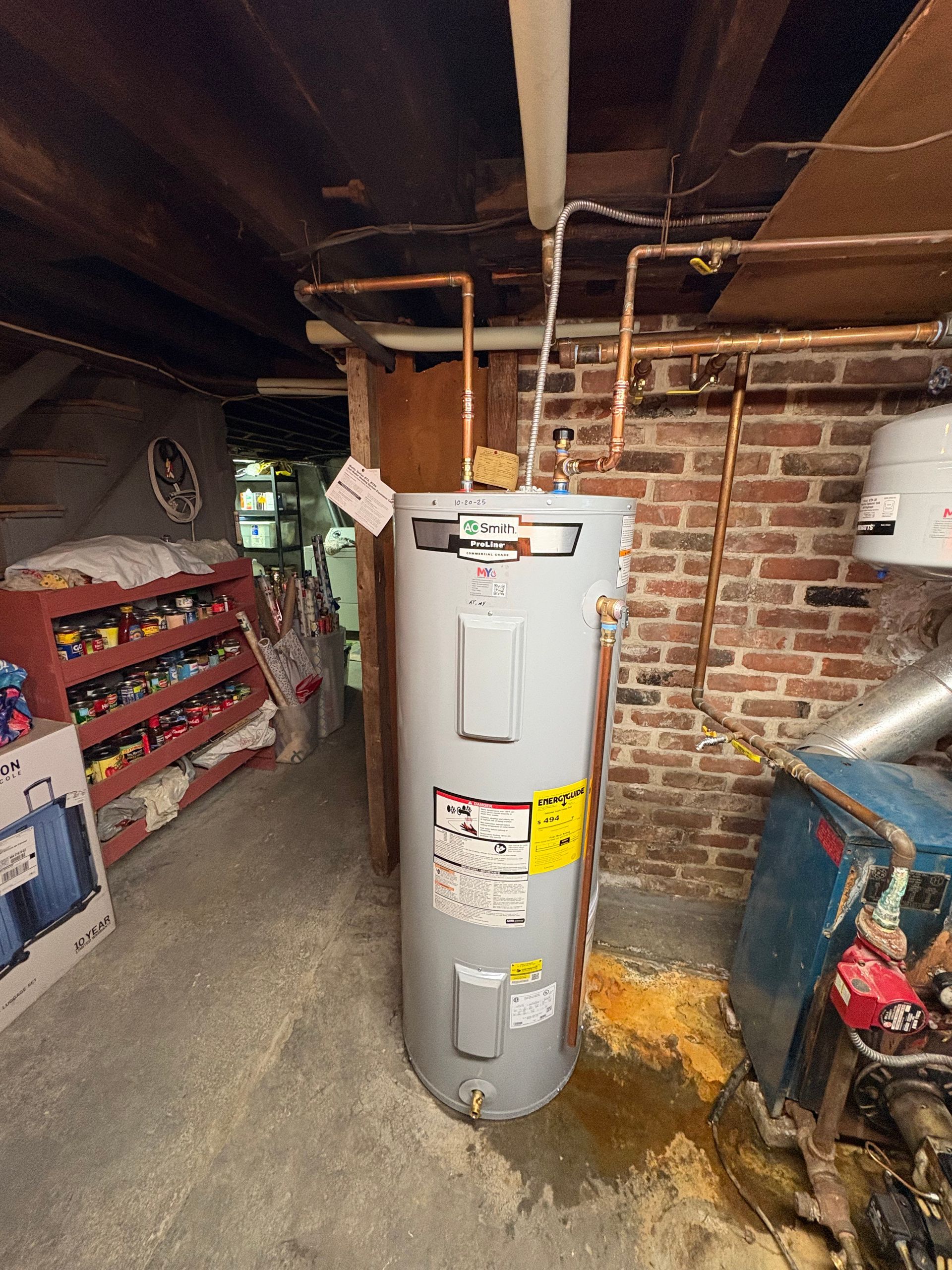 Water heater in a basement with brick walls, copper pipes, and a concrete floor.