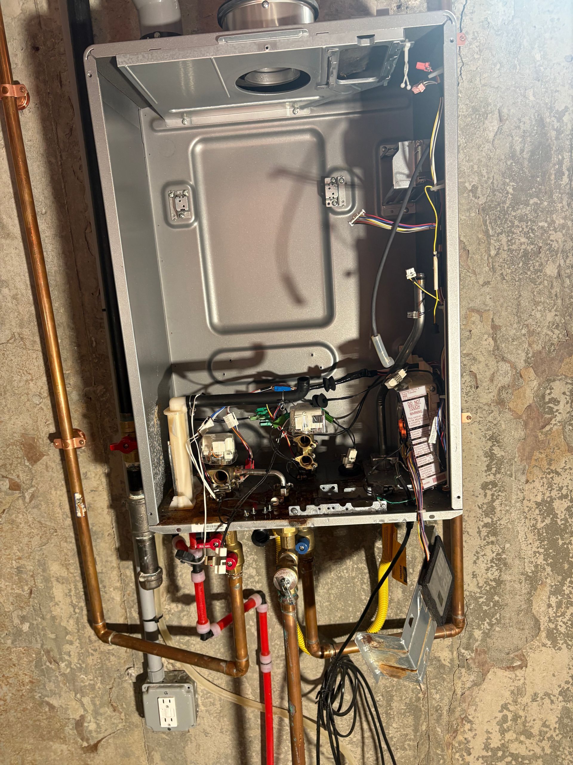 Opened wall-mounted boiler showing internal components and copper piping on a concrete wall.