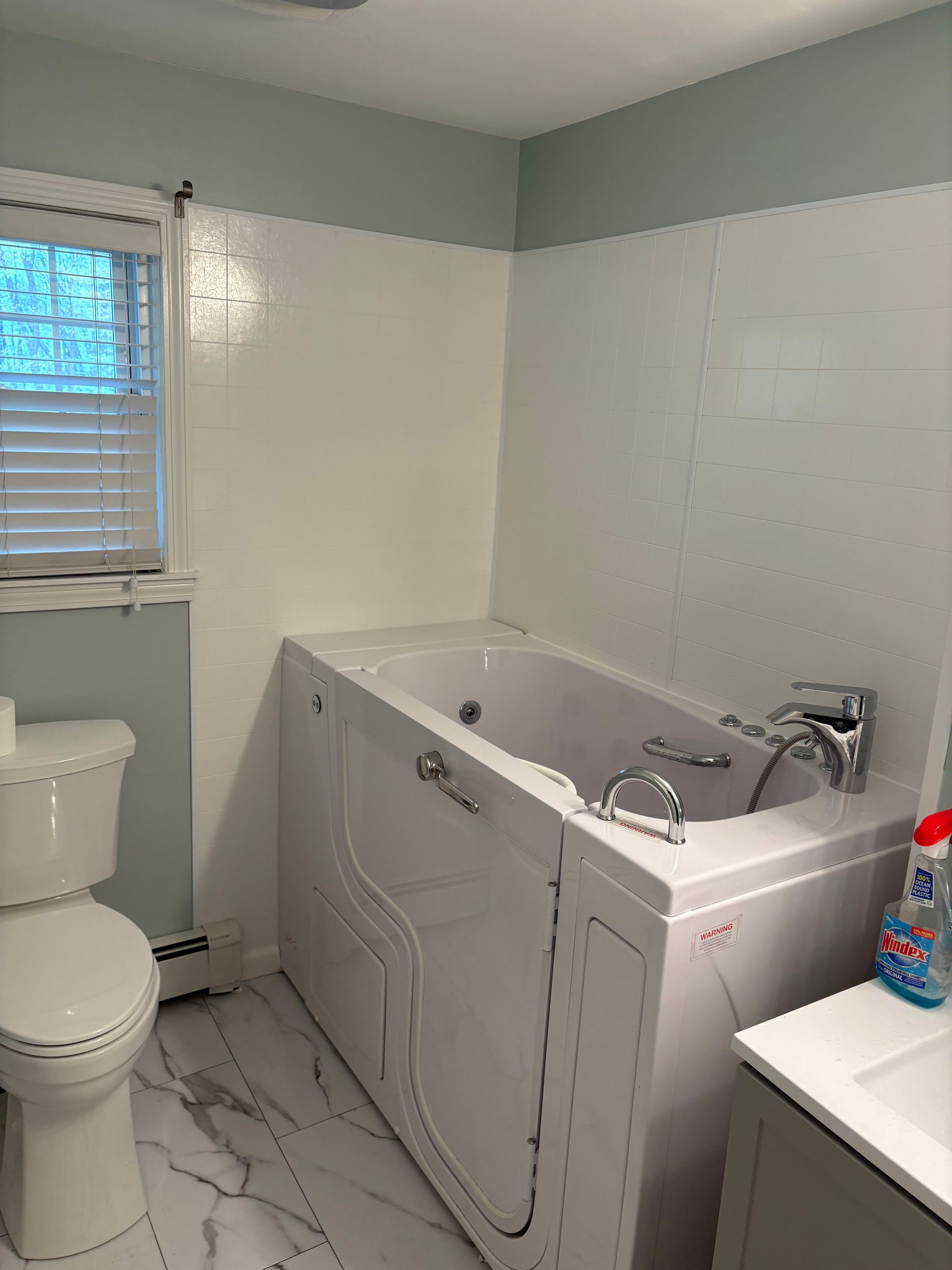 Bathroom with walk-in tub, toilet, and vanity. White and pale blue walls, window with blinds, marble-look floor.