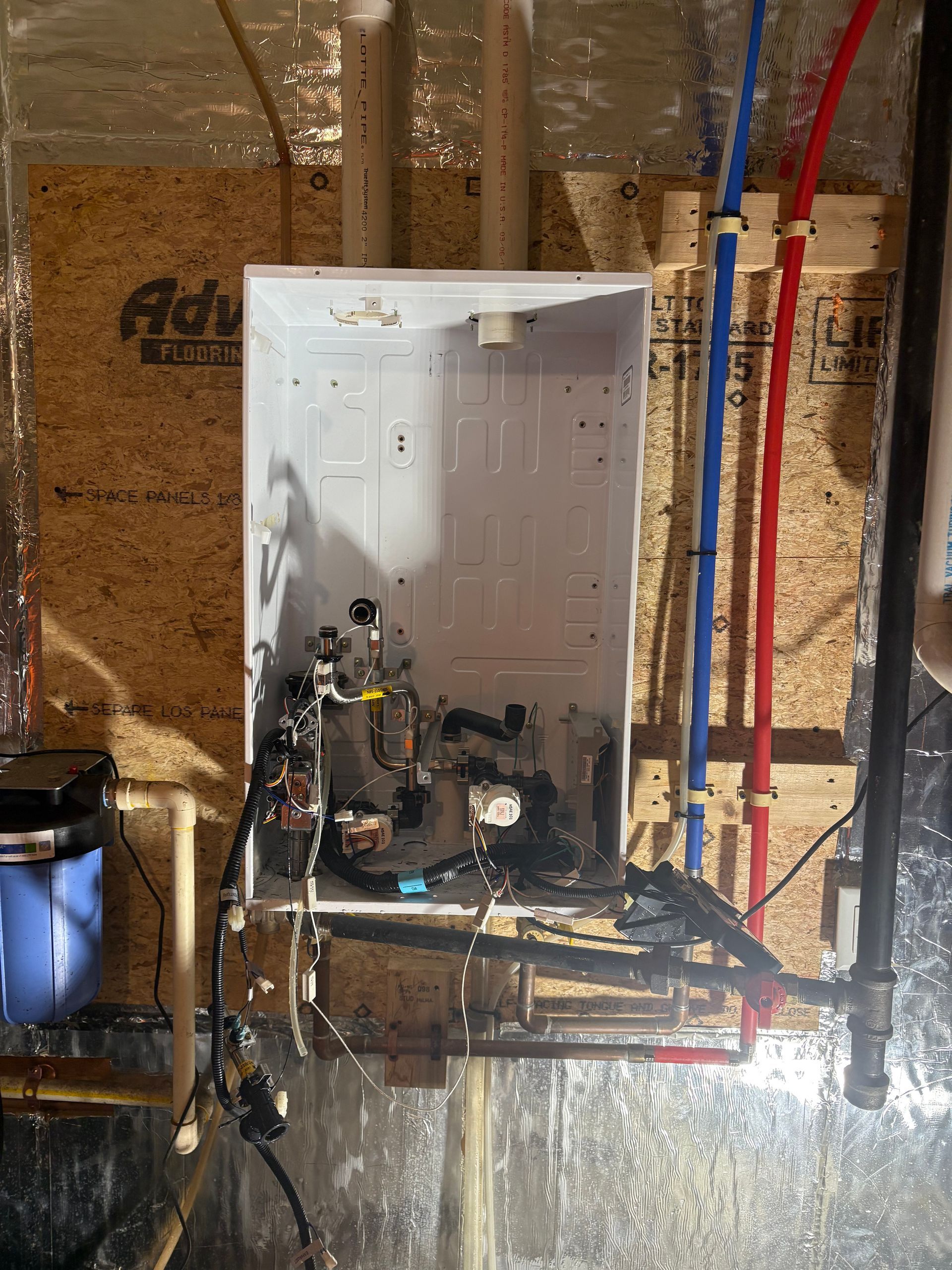 White tankless water heater with exposed internal components, mounted on plywood. Blue and red PEX pipes visible.