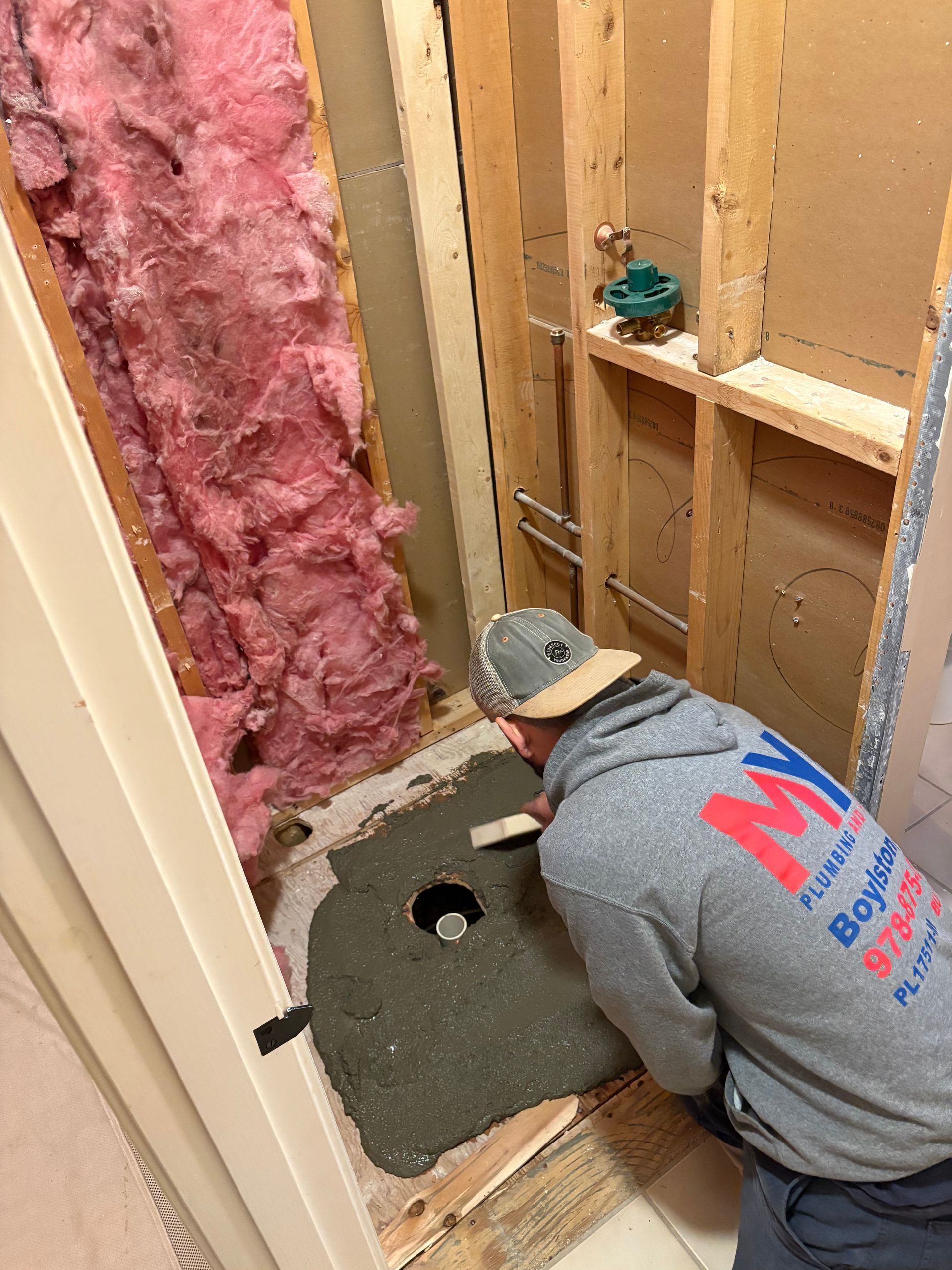 Man applying cement to a shower floor. Construction site. Pink insulation and wood framing visible.