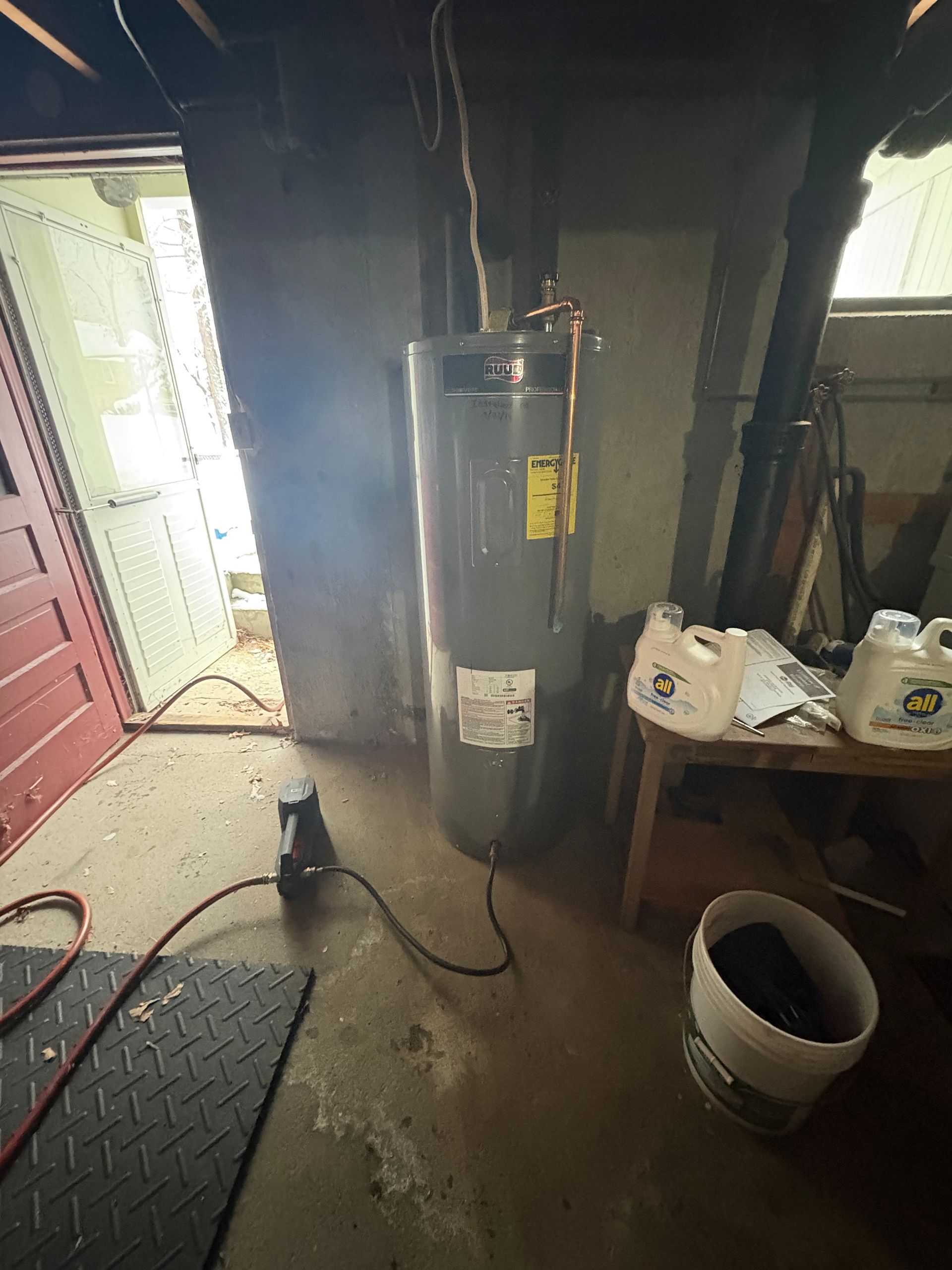Water heater in a basement next to an open doorway.  A bucket and containers are nearby.