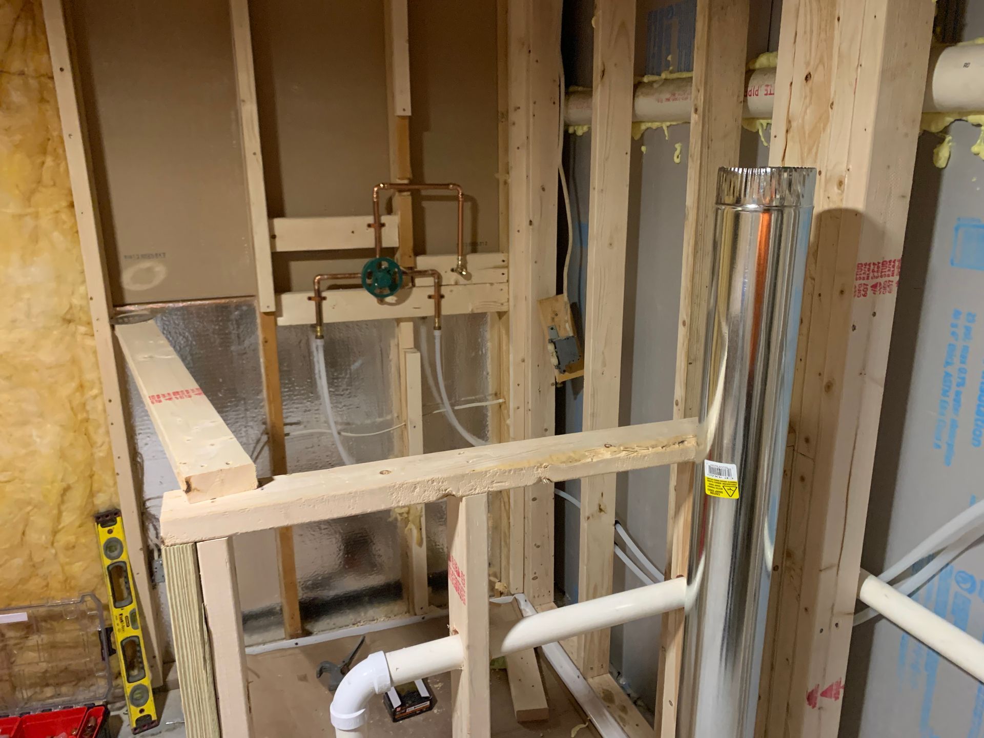 Framing of a wall with exposed copper plumbing, a silver flue, and white PVC pipes in a construction setting.