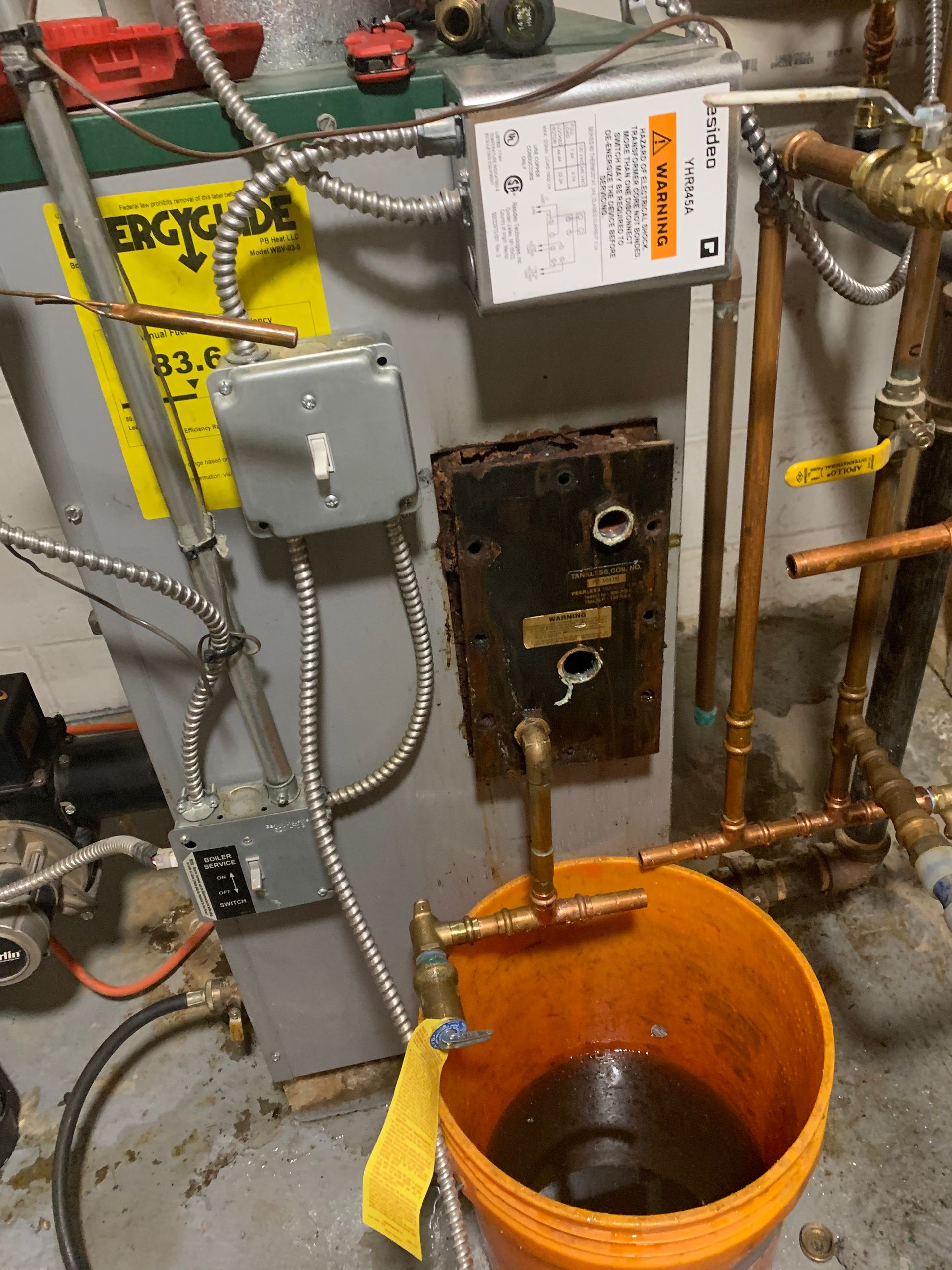A boiler with exposed components, a bucket catching water, and various copper pipes.