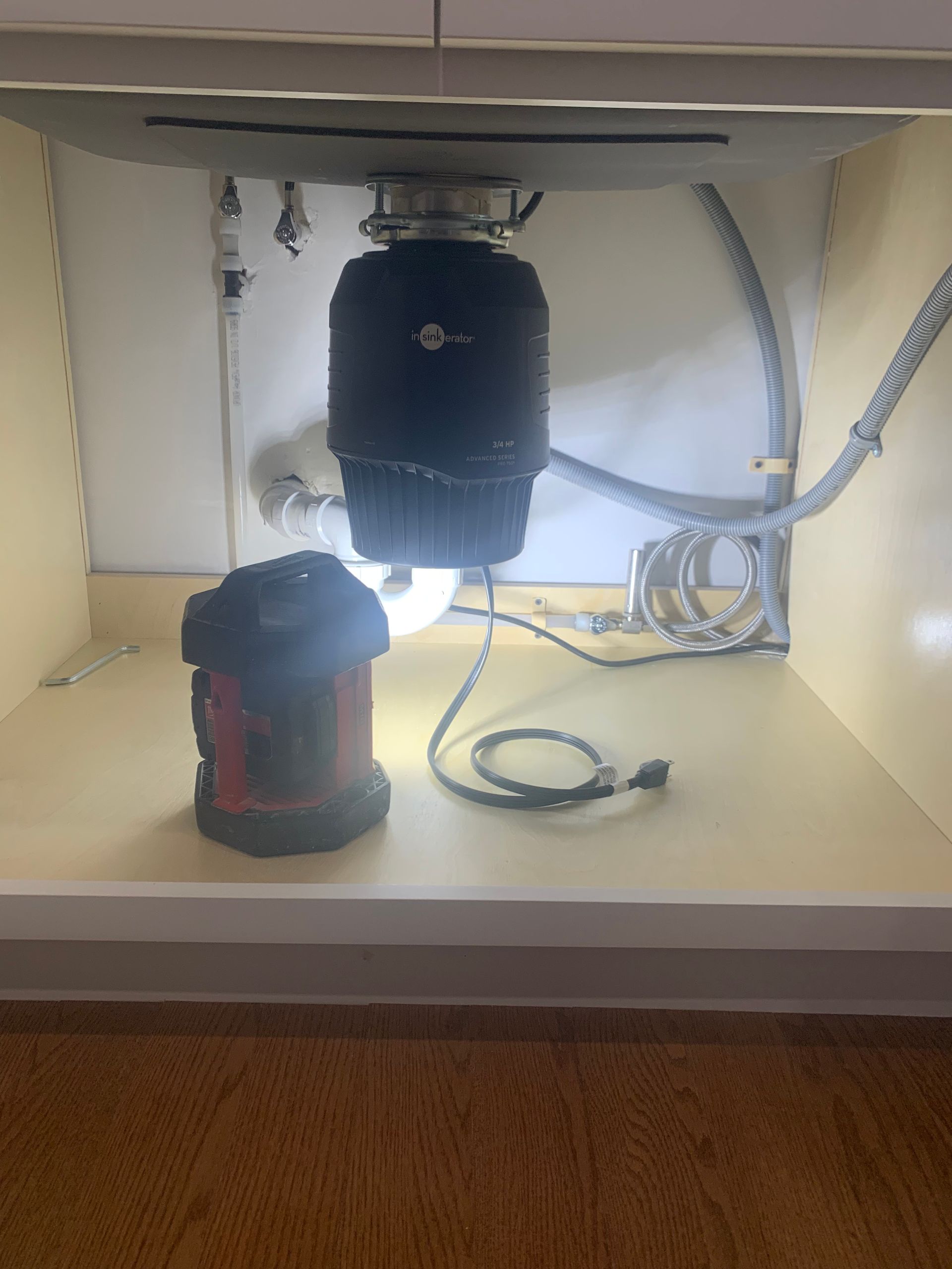 Under-sink view with a garbage disposal unit, water pipes, and a red work light.