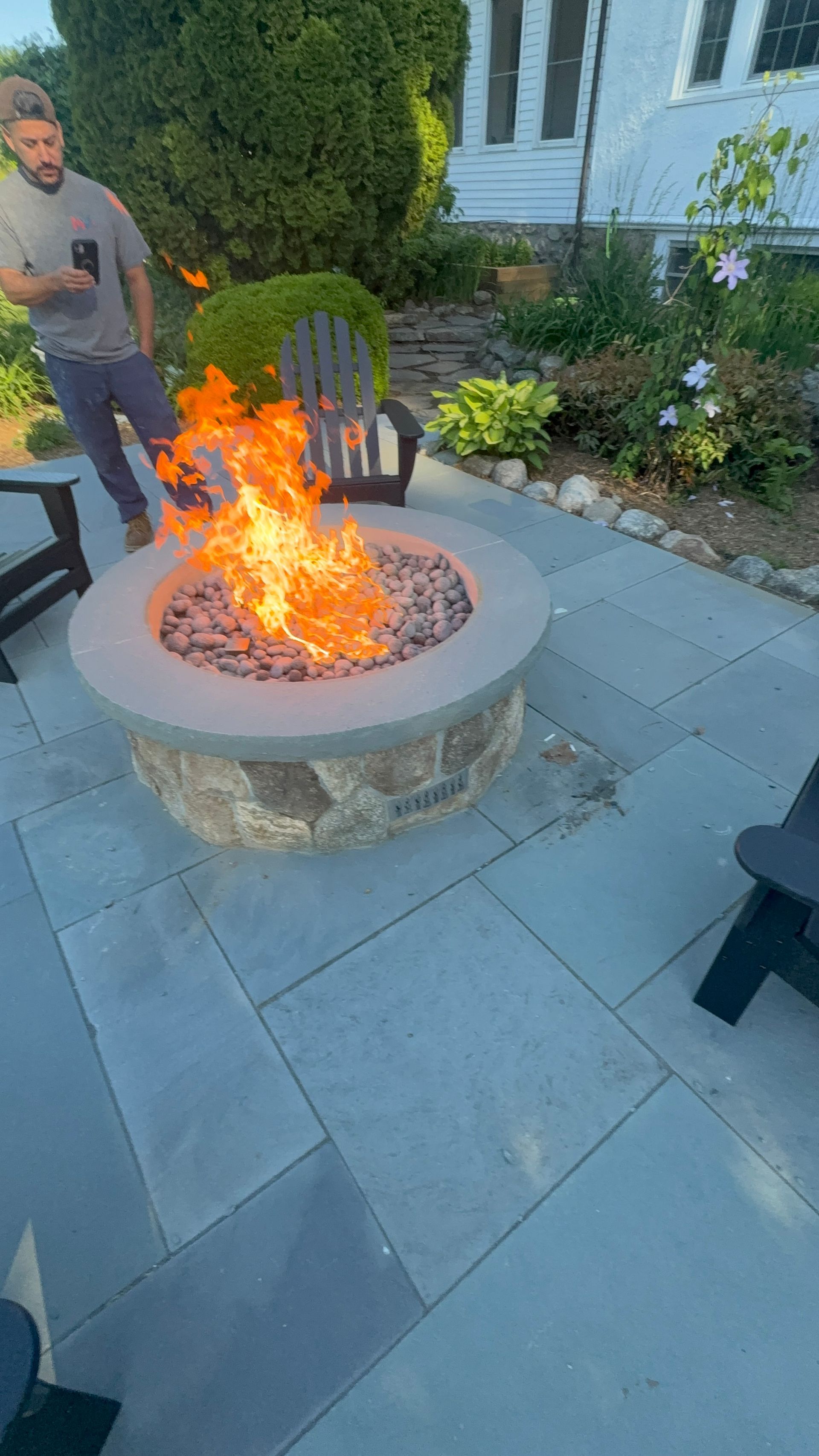 Backyard stone fire pit burning on a patio, with a person standing nearby and chairs around it