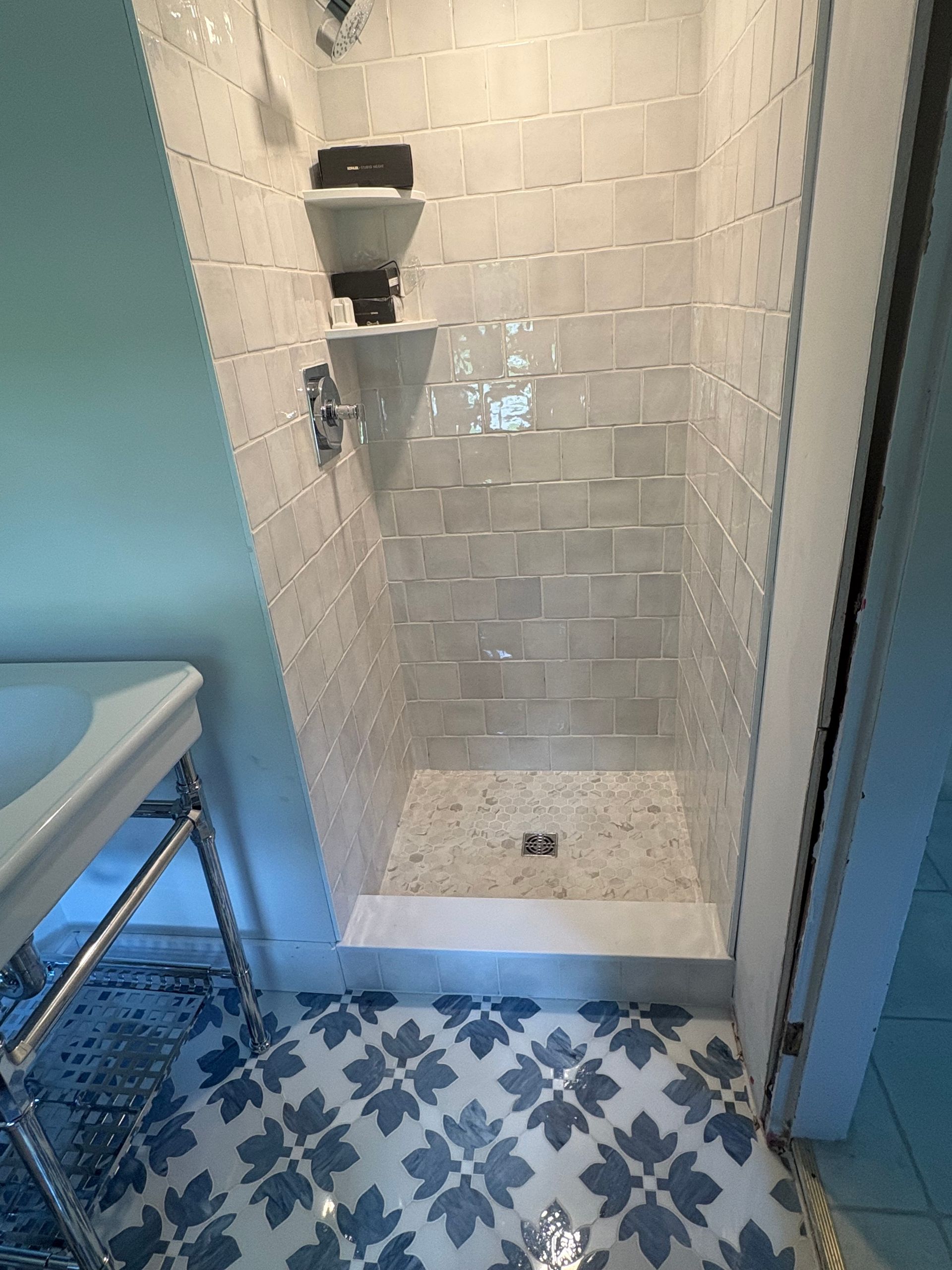 Small tiled shower stall with built-in shelf, next to a blue padded bench and patterned blue-and-white floor.