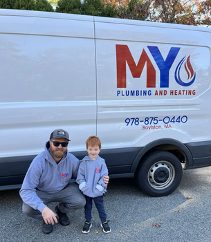 A man and child kneel beside a MY Plumbing and Heating van on a gravel lot.