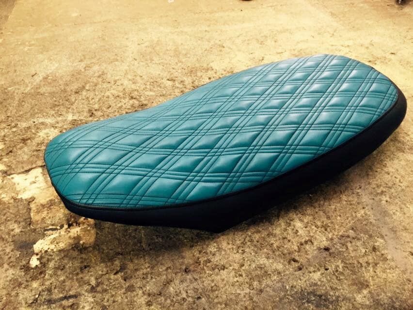 Teal diamond-stitched motorcycle seat with black trim, resting on a textured surface.