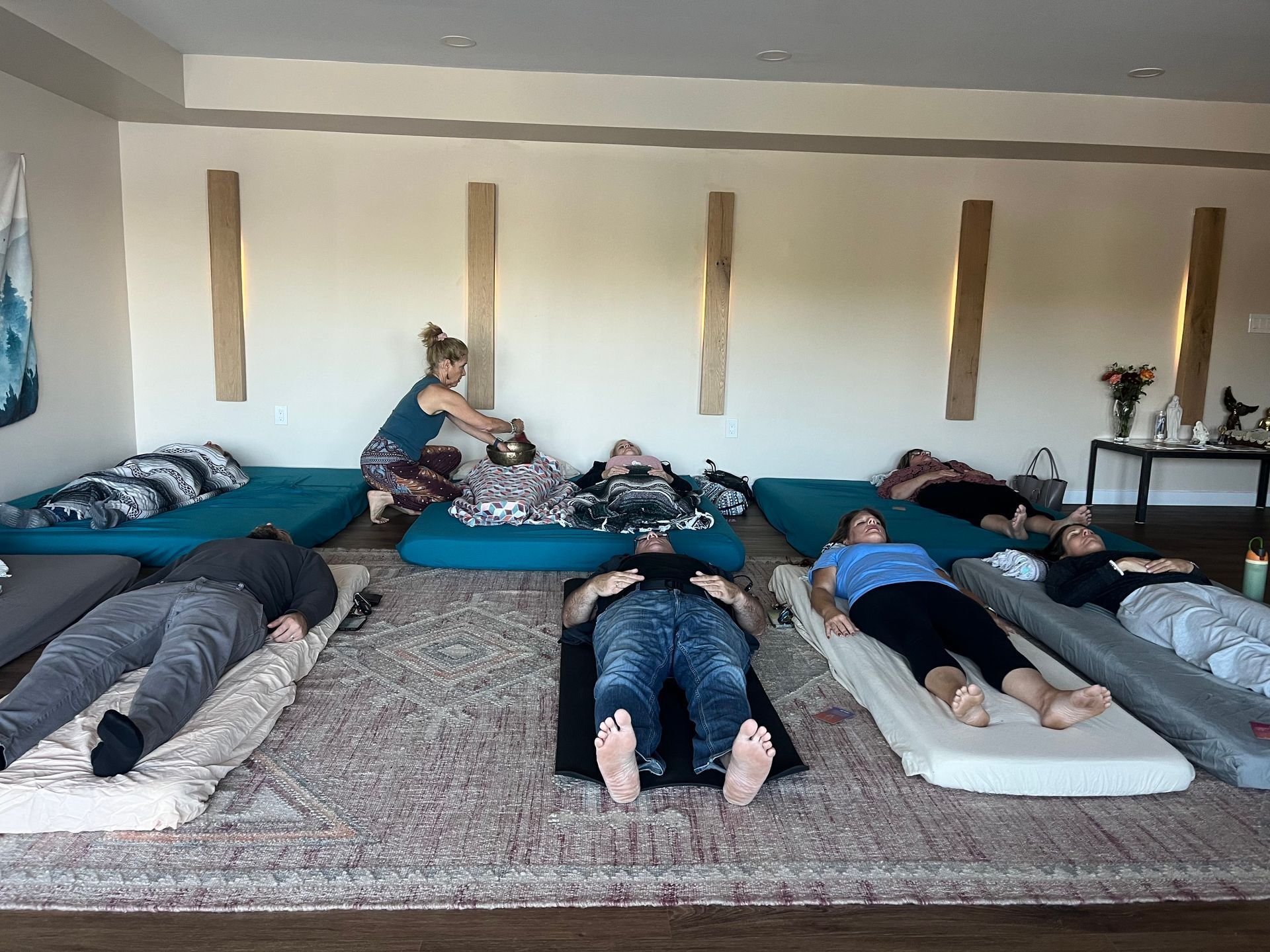 People lying on mats in a room; a woman adjusts one person’s blanket. Sunlight streams in.