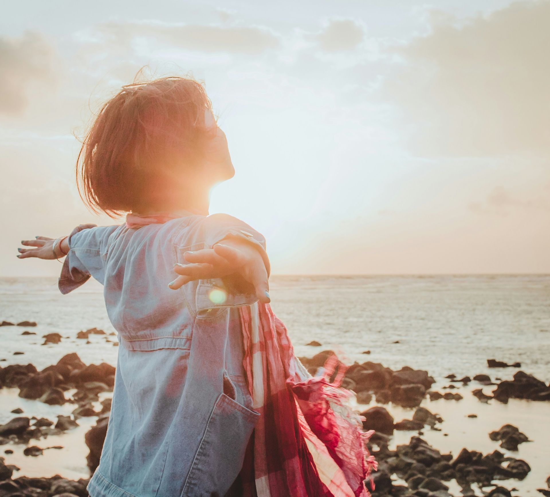 Woman with arms outstretched, facing sun over ocean. Rocks in foreground, warm light.