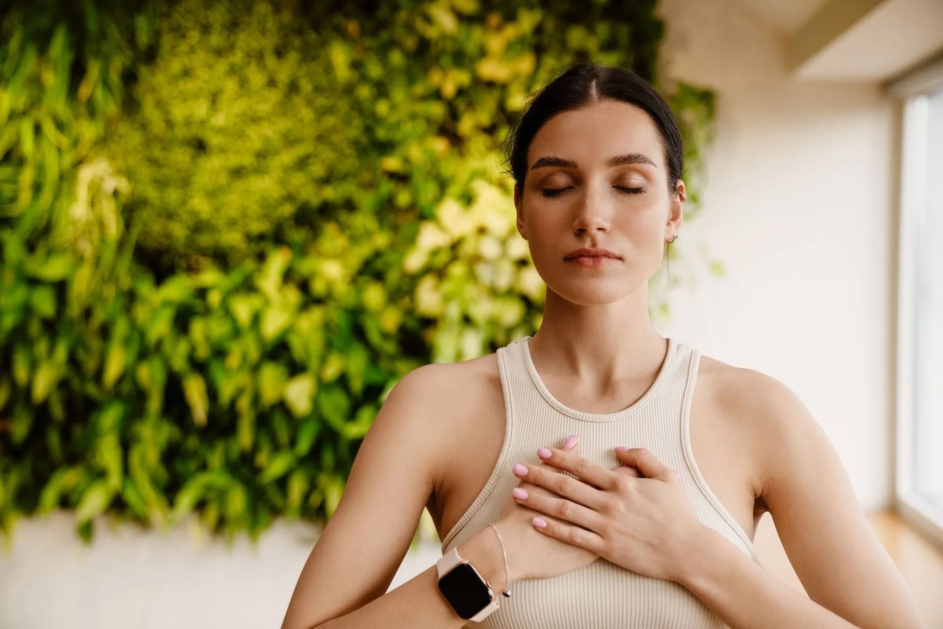 Woman meditating, eyes closed, hands on chest, in front of a green plant wall.