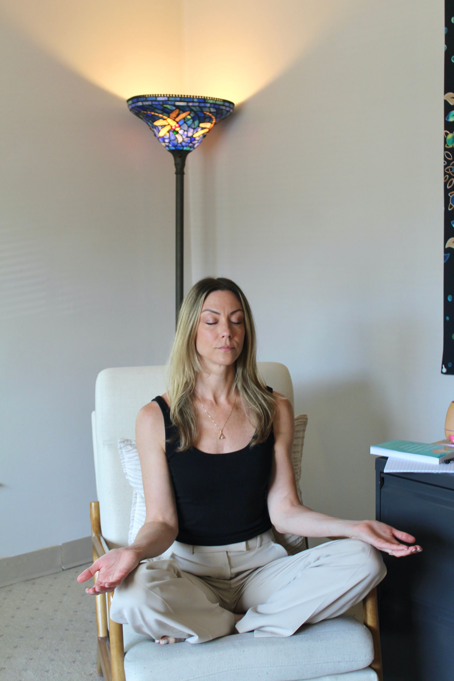 Woman meditating, seated in a chair, eyes closed, hands open. Light from a stained-glass lamp above.