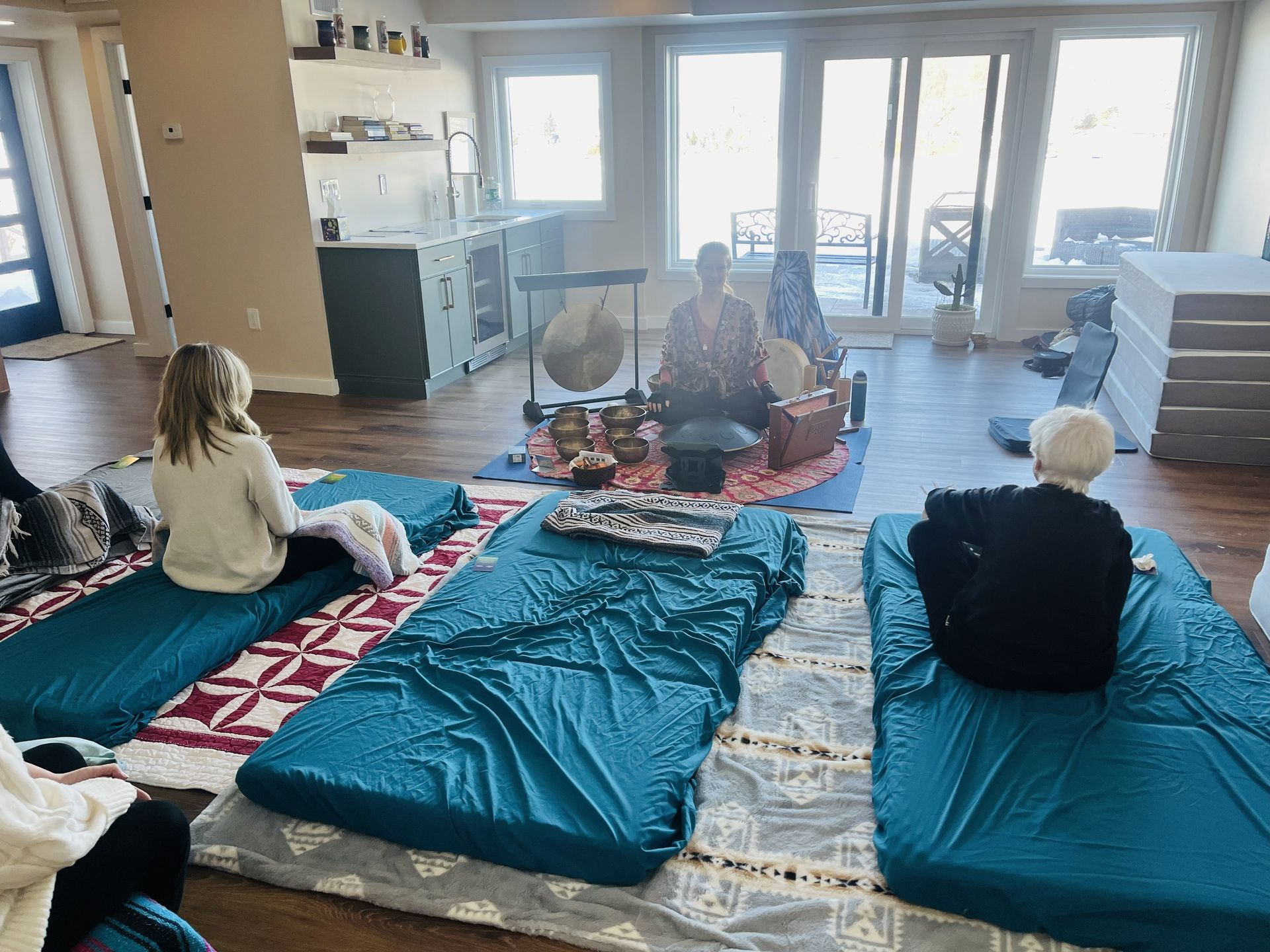 Three people sit on floor mats in a bright room, facing a leader behind a gong and singing bowls, suggesting a sound bath.
