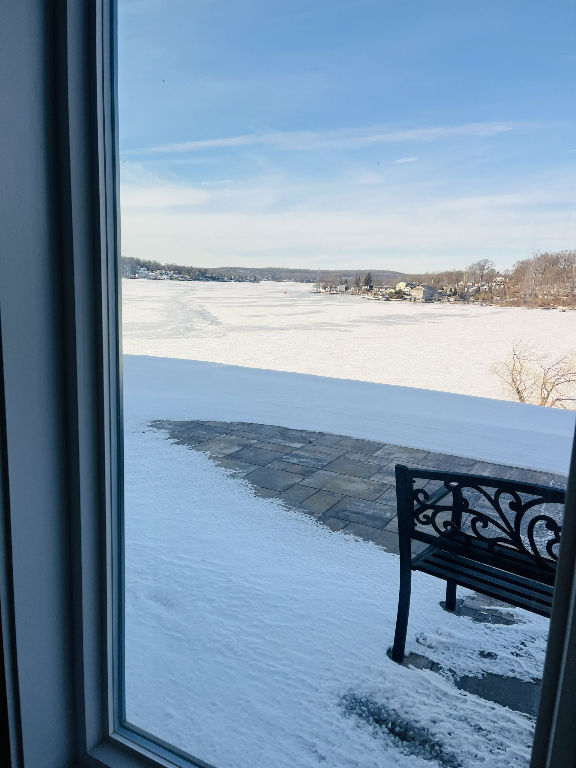 A view through a window overlooking a snow-covered patio and a frozen, ice-covered lake under a clear blue sky.