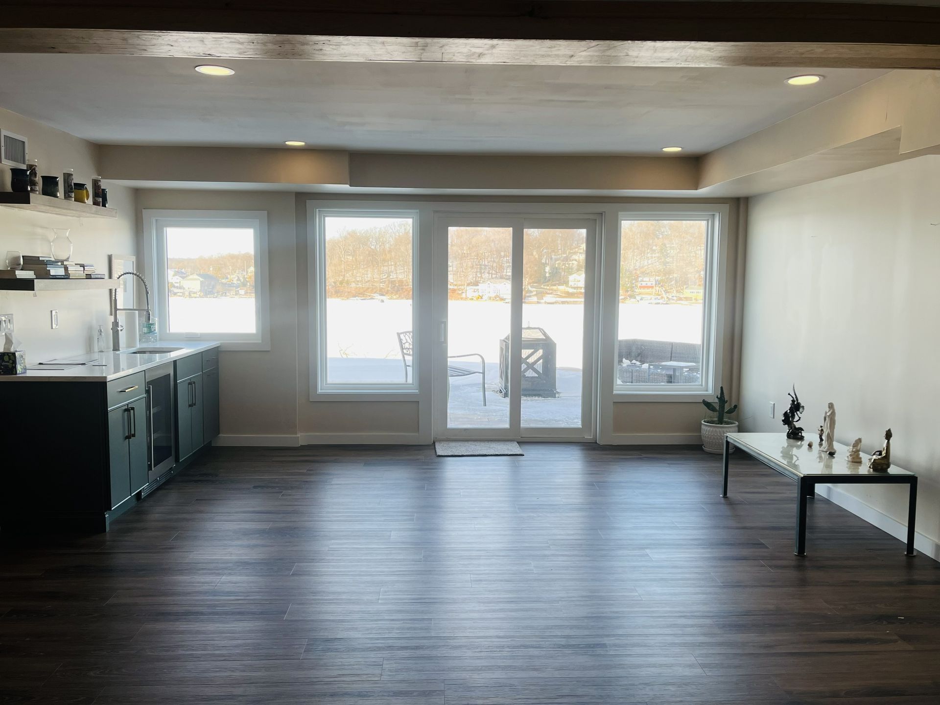 A modern room with dark wood floors, a kitchen counter on the left, sliding glass doors to a snowy yard, and a side table.