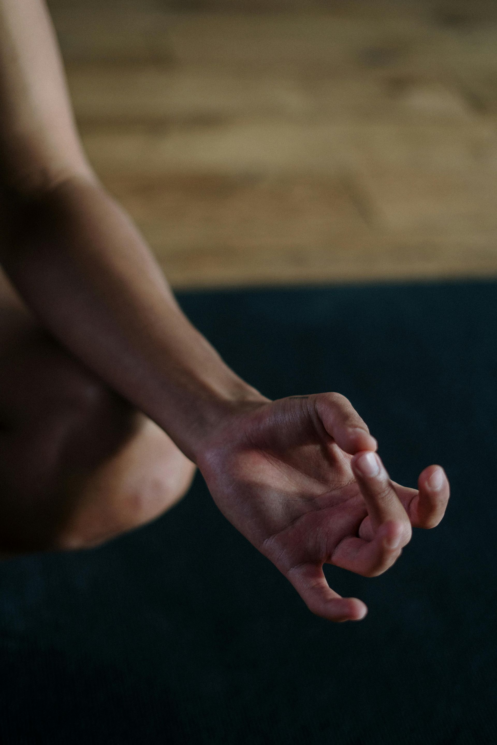 Hand with fingers curled in a yoga mudra, near a dark mat and wood floor.