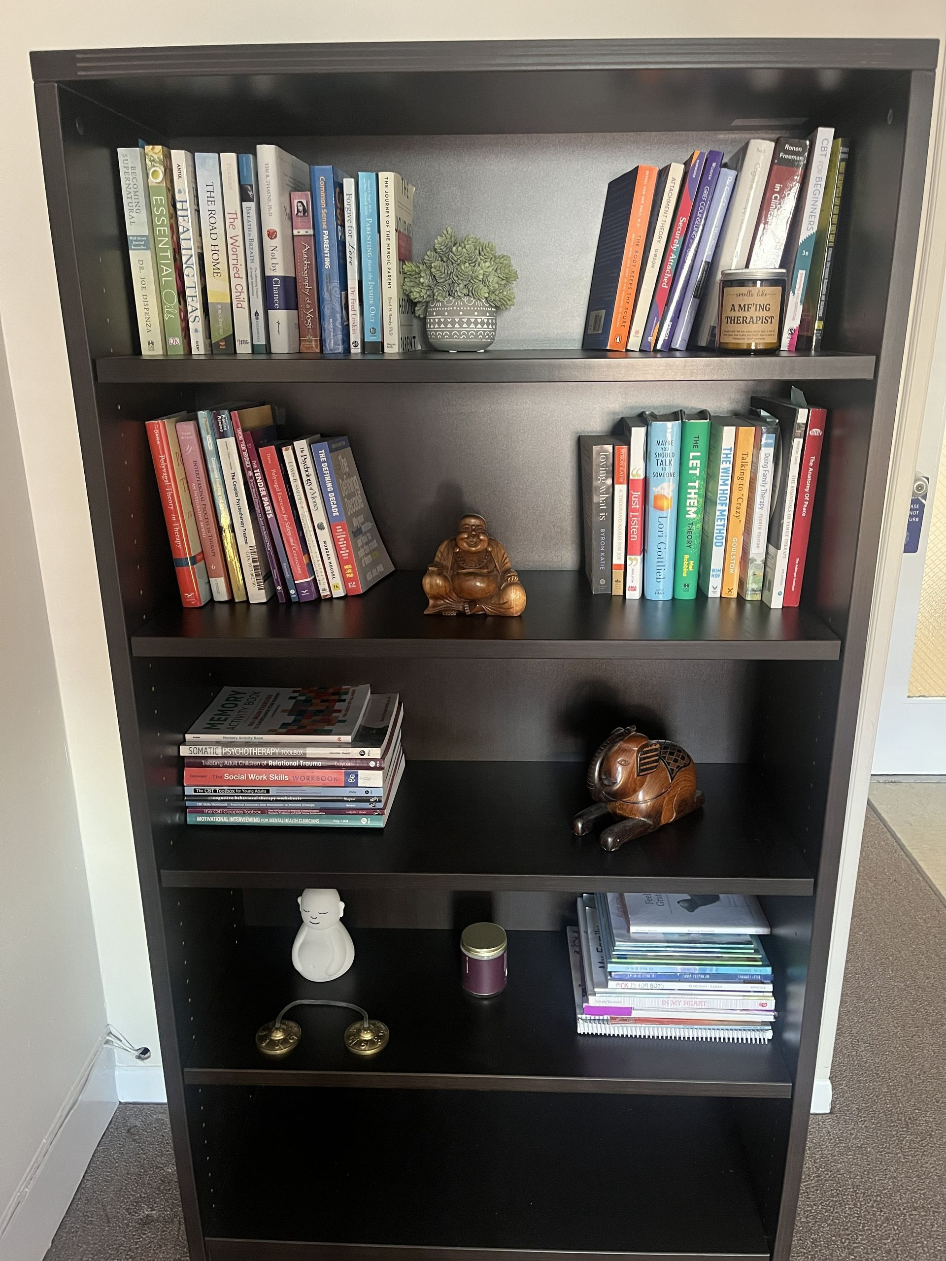 A dark brown wooden bookshelf with five shelves, containing various books, small decorative items, and a Buddha figurine.