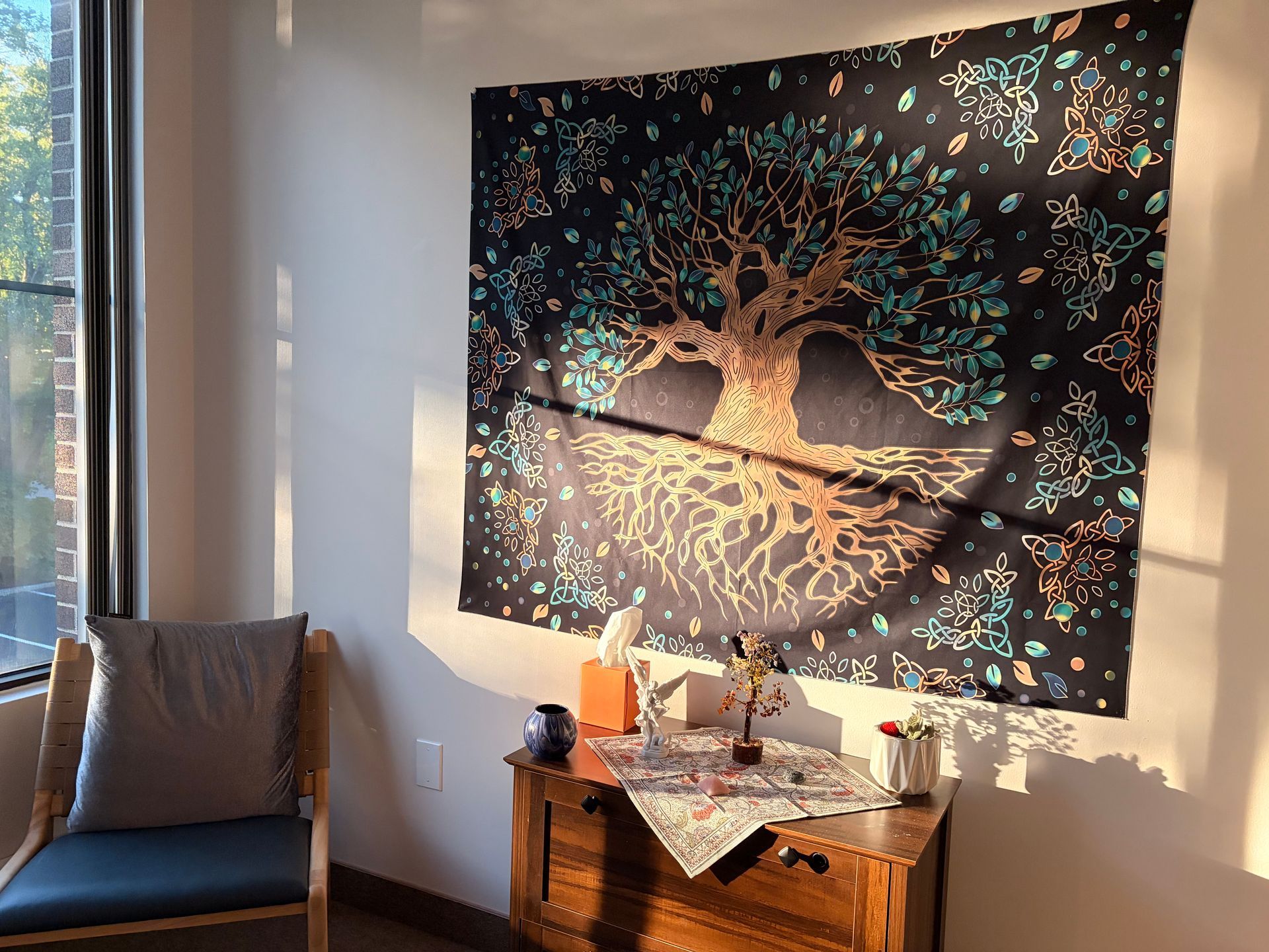 Sunlit room with a tree tapestry above a small wooden table and chair by the window