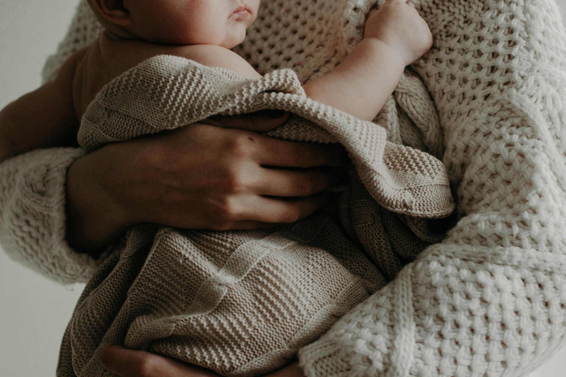 A person holding a swaddled baby; soft tan blanket and cream sweater are visible.