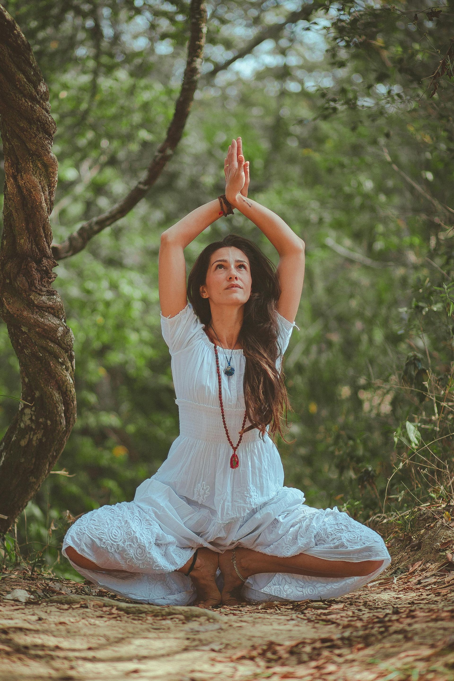 Woman in white dress meditating outdoors, hands raised, eyes closed, surrounded by trees.