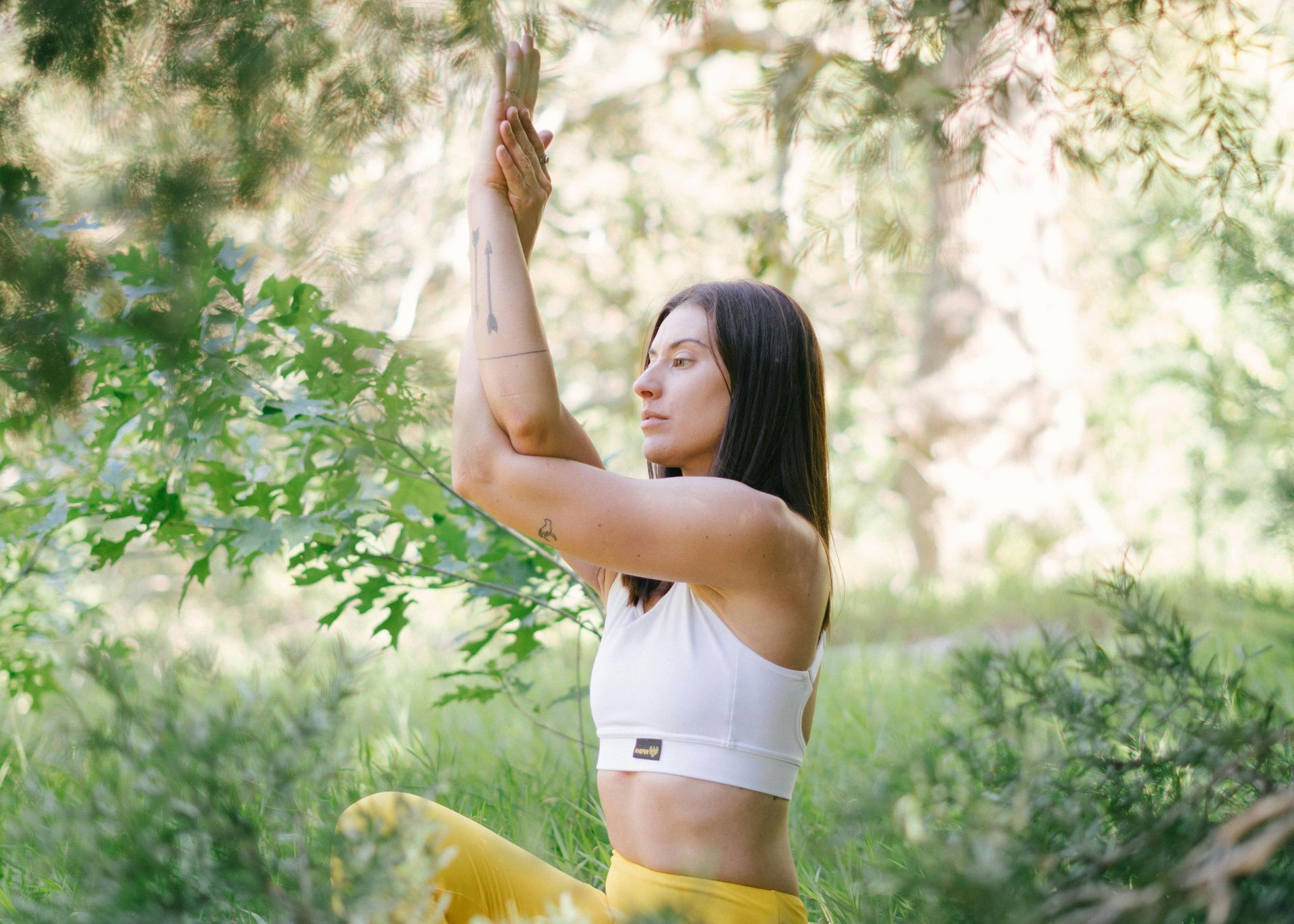 Woman in white top, yellow pants, practicing yoga pose outdoors. Arms crossed, eyes closed.
