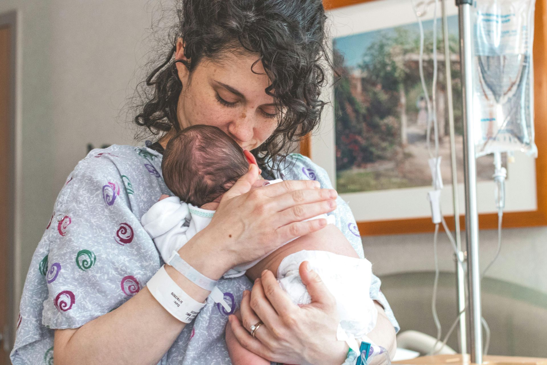 Woman in hospital gown kisses newborn baby's head; saline drip in background.