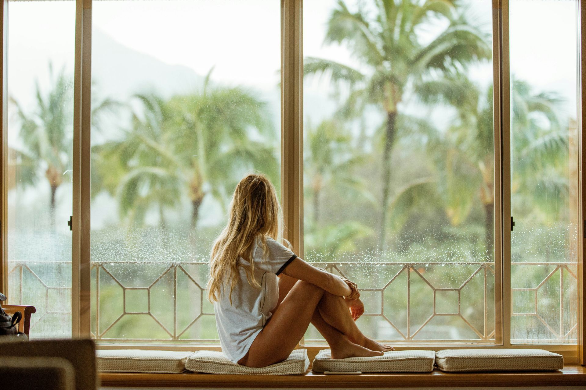 Woman sitting by a window, looking out at palm trees. Natural light, relaxed pose.
