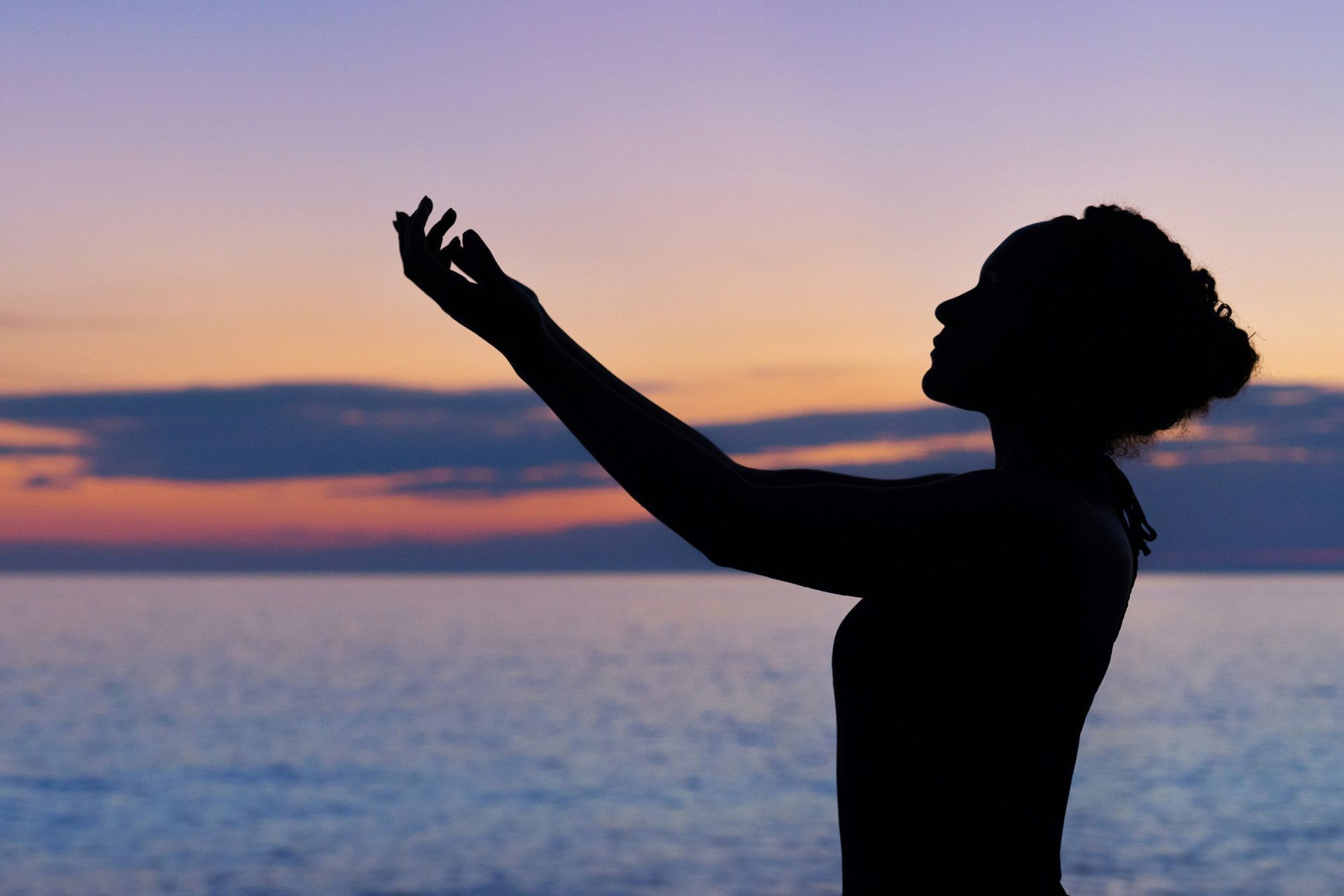 Silhouette of a person with arms outstretched, facing sunset over water.