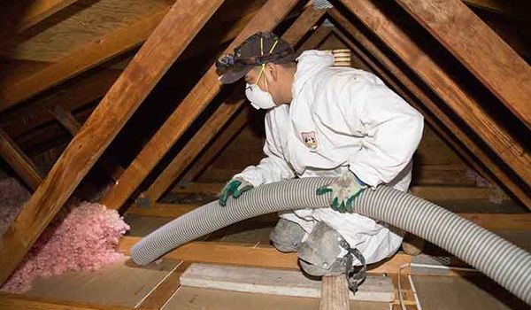 Worker in white protective suit spraying insulation into an attic with exposed wooden beams.