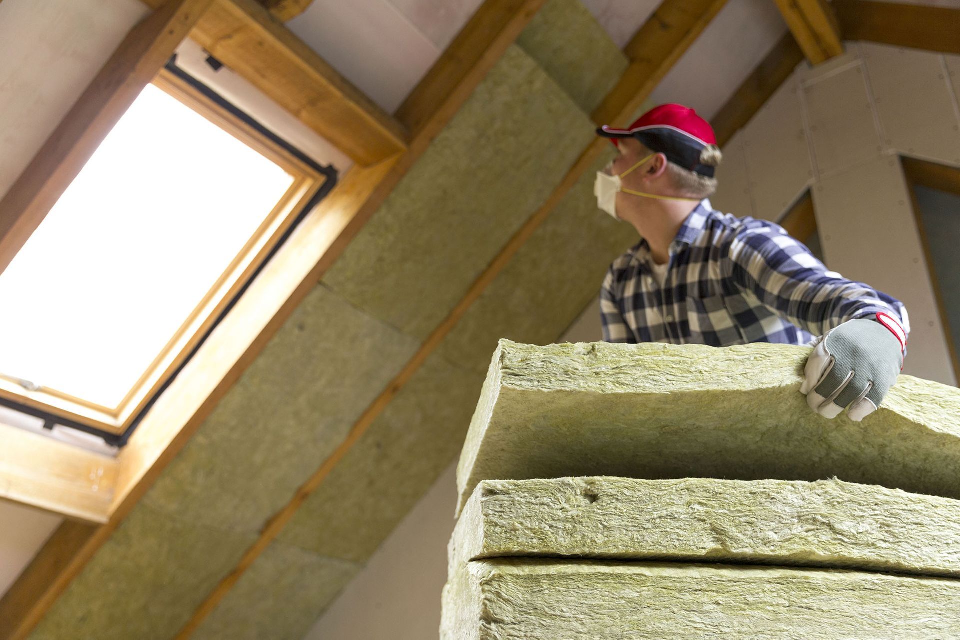 Worker in red cap stacking insulation panels in an attic by a skylight.