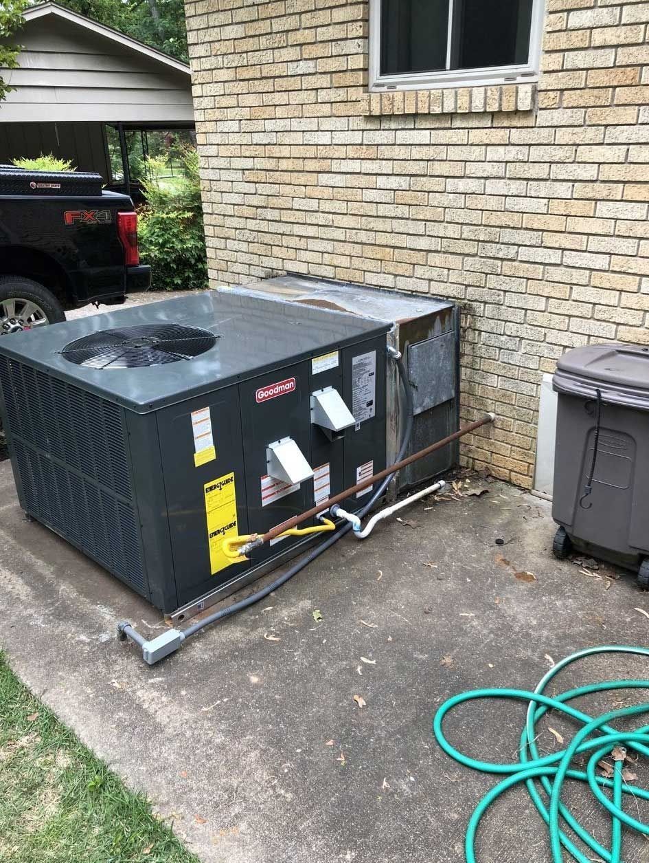 Air conditioning unit next to a brick wall and trash can on a concrete slab. Green hose on ground.