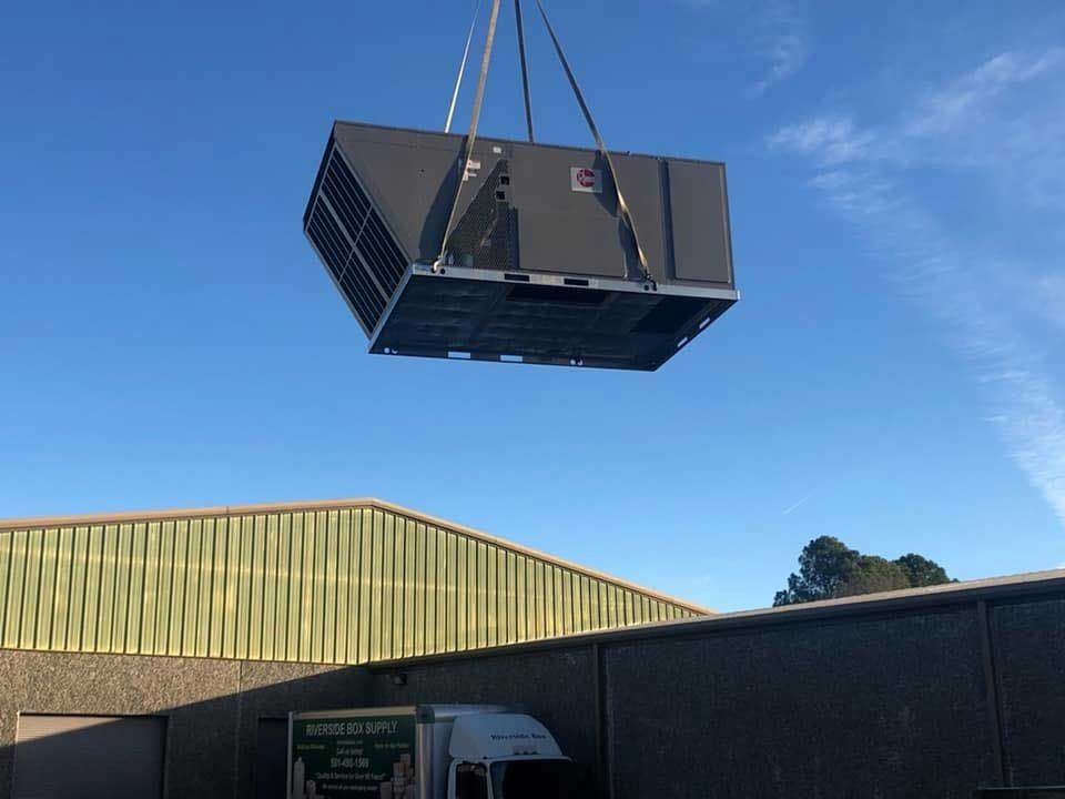 A large HVAC unit is lifted by a crane against a blue sky above a building.