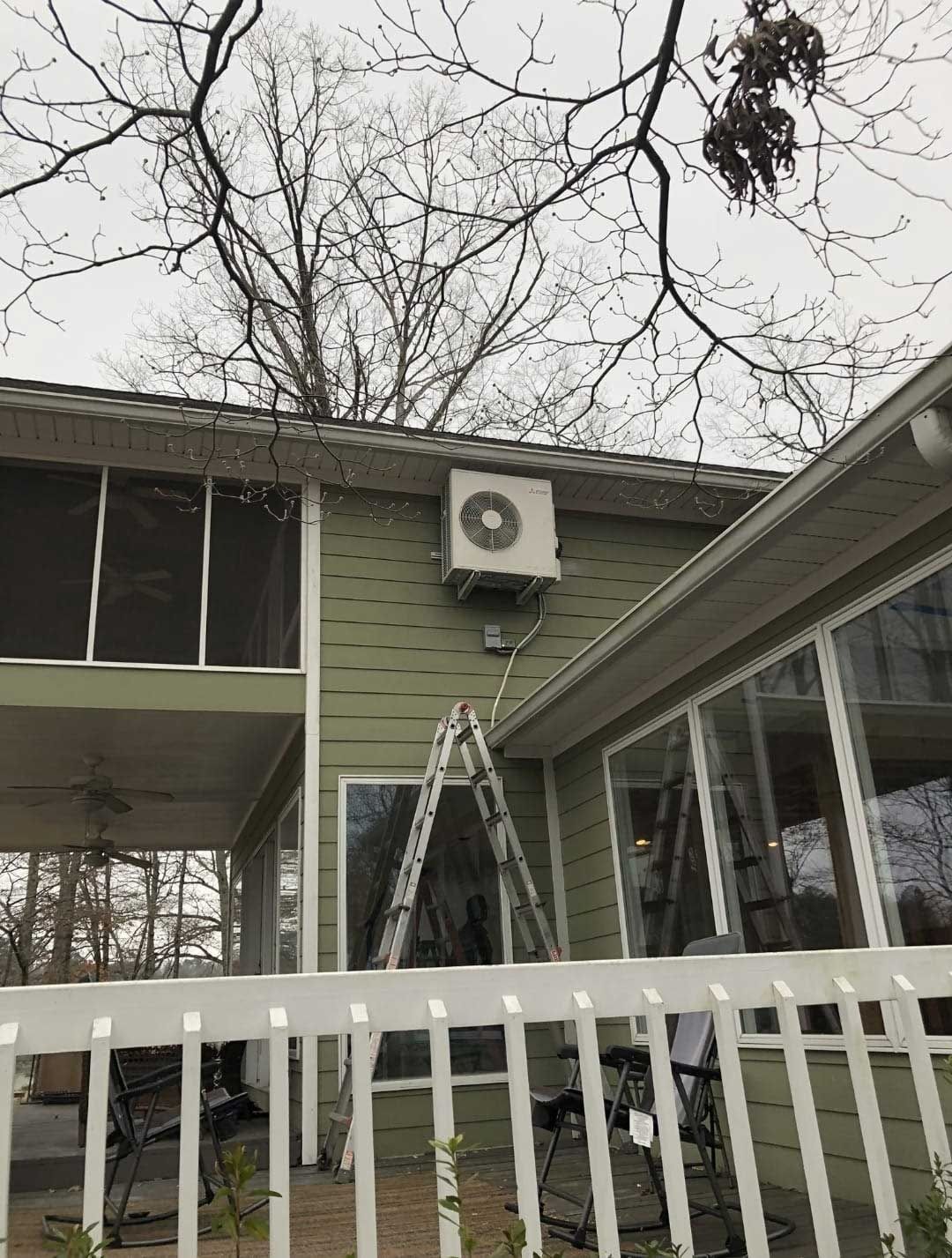Outdoor wall-mounted air conditioner on a green house exterior, with a ladder and trees in the background.