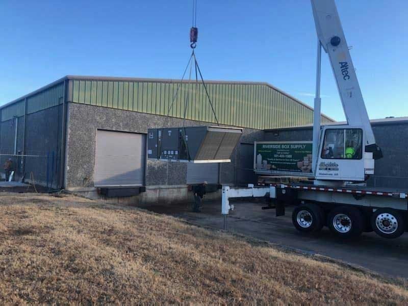 A crane lifting an HVAC unit near a building. The sky is blue, and there is dry grass in front.