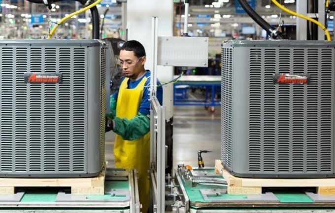 Factory worker inspects air conditioners on an assembly line; person in yellow apron and green gloves.