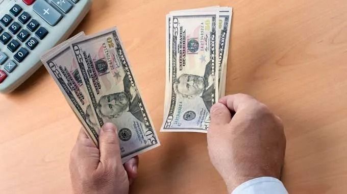 Hands counting US fifty-dollar bills on a wooden surface, a calculator visible in the background.