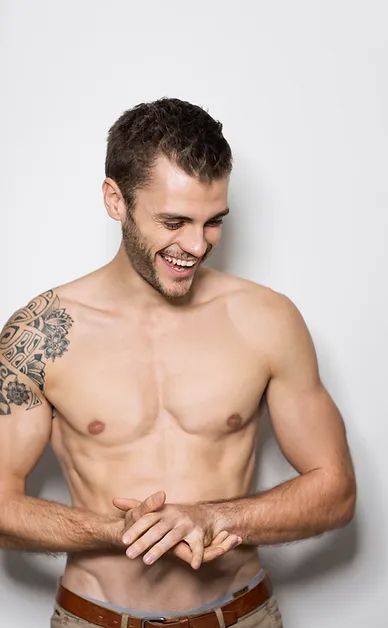 A shirtless man with a tattoo on his shoulder is smiling.