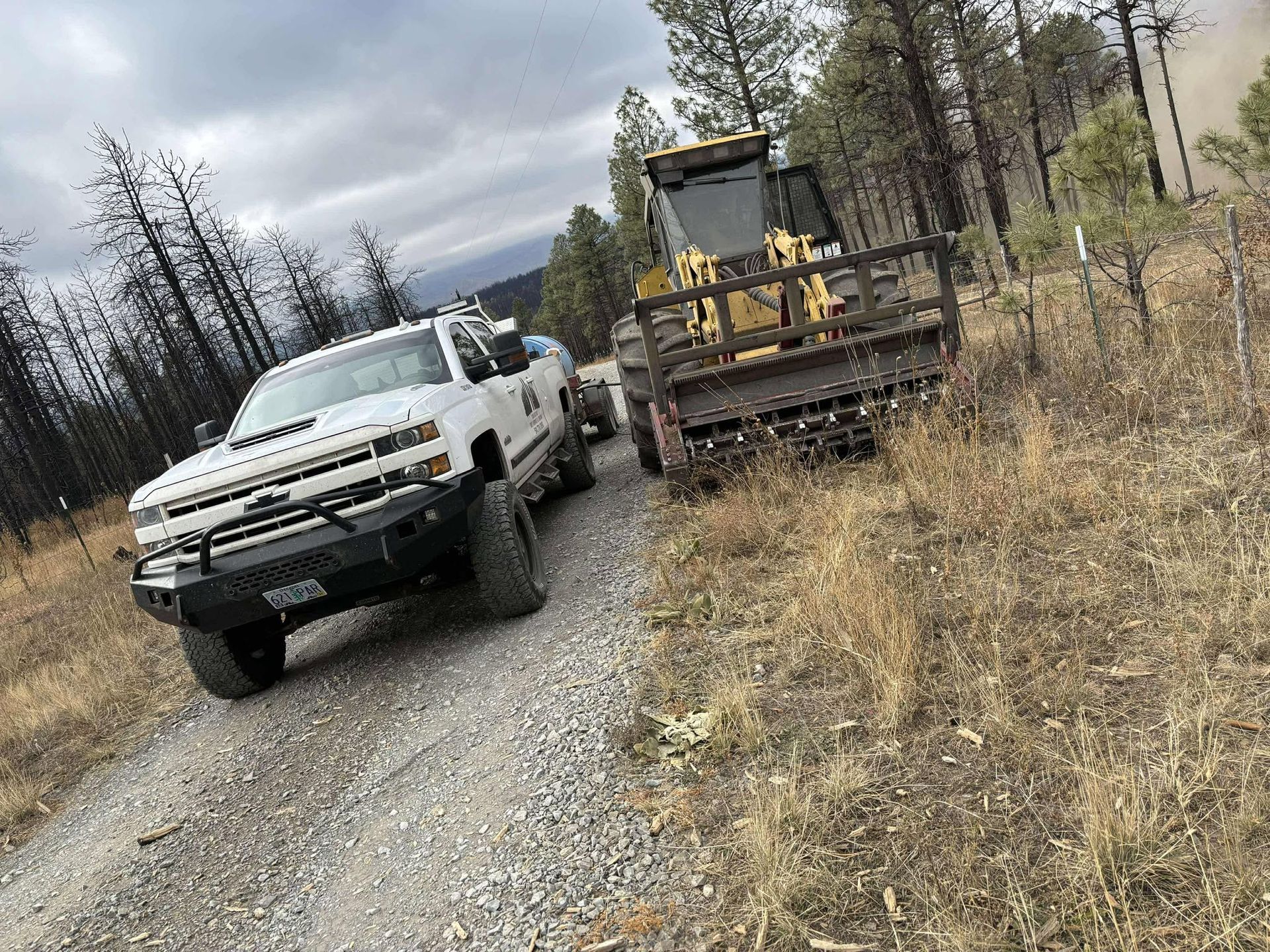 White pickup truck towing a heavy machine on a dirt road in a wooded area, cloudy sky.