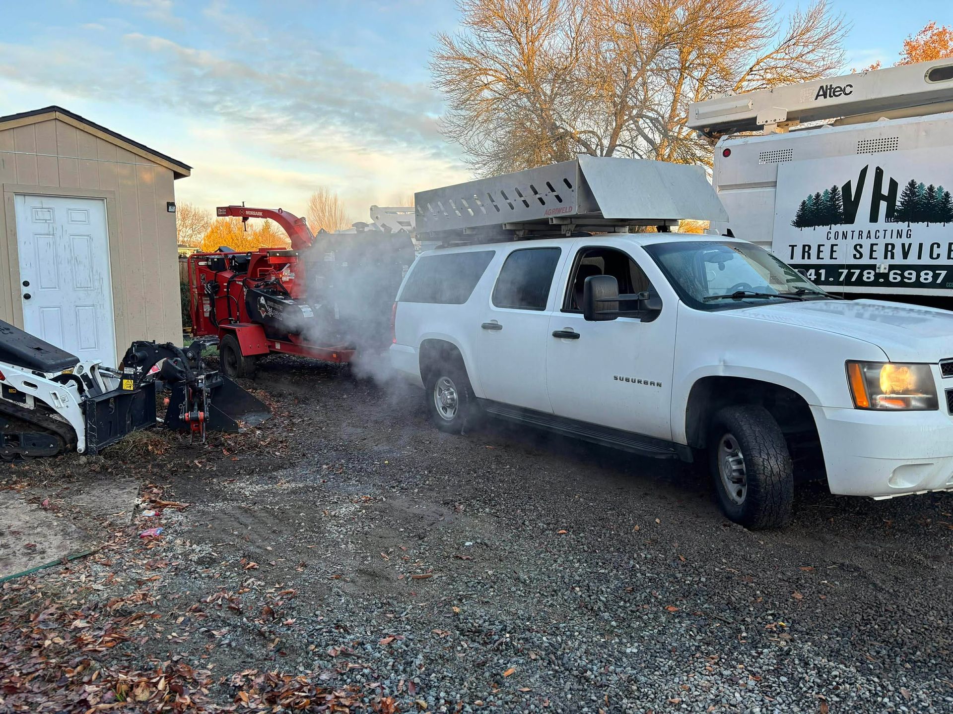 A white SUV with a roof rack and a wood chipper are parked near a shed. Smoke rises from the chipper.