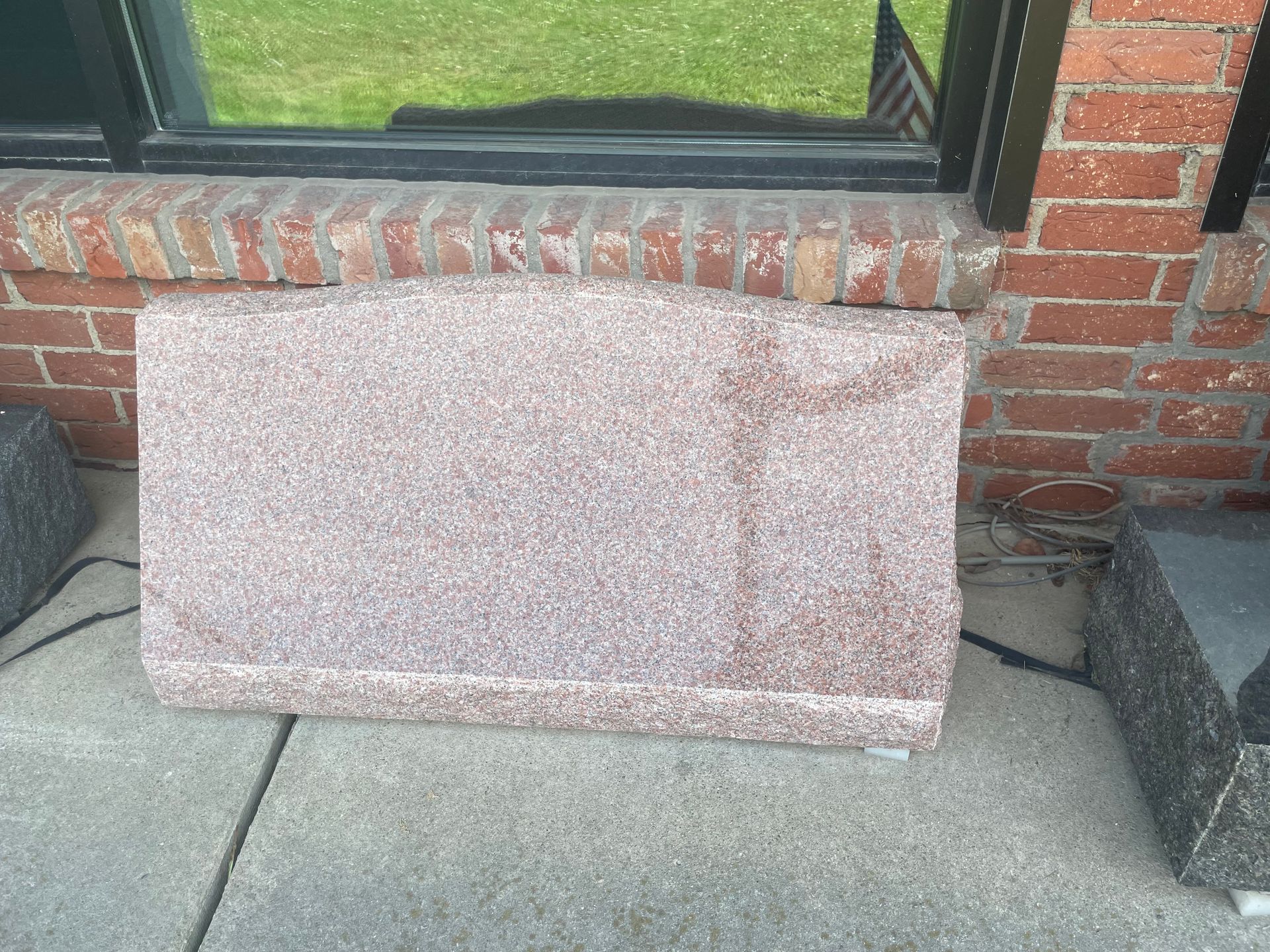 A rectangular, speckled pink-toned object rests on a concrete surface, positioned below a window and brick wall.