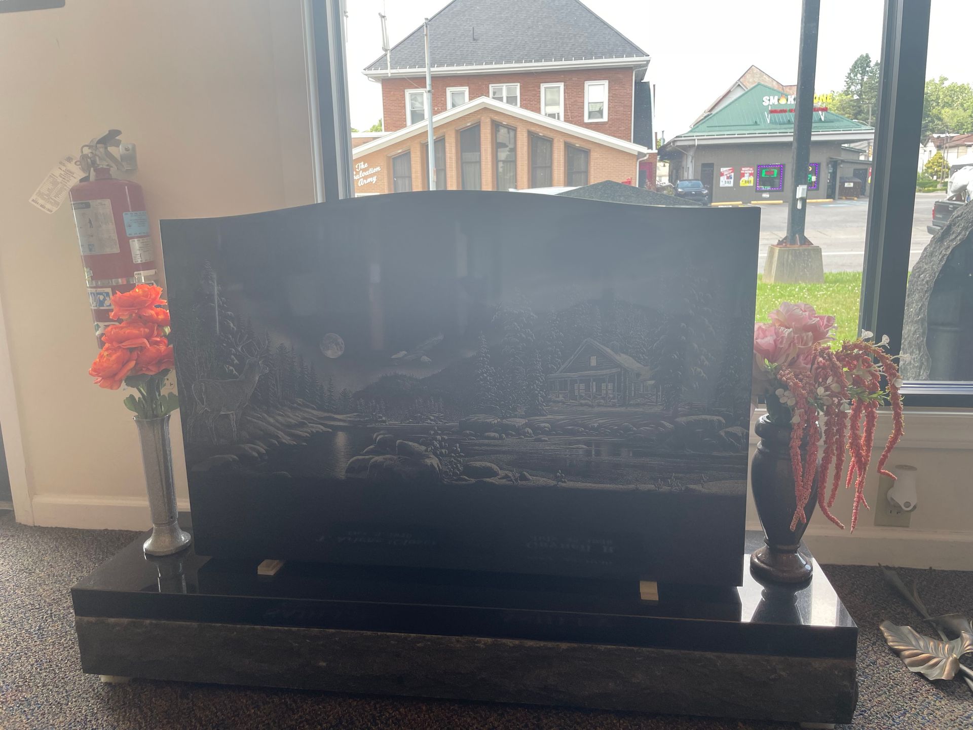 Black granite headstone with an engraved scene, flanked by vases of flowers, inside a building with a window showing buildings.